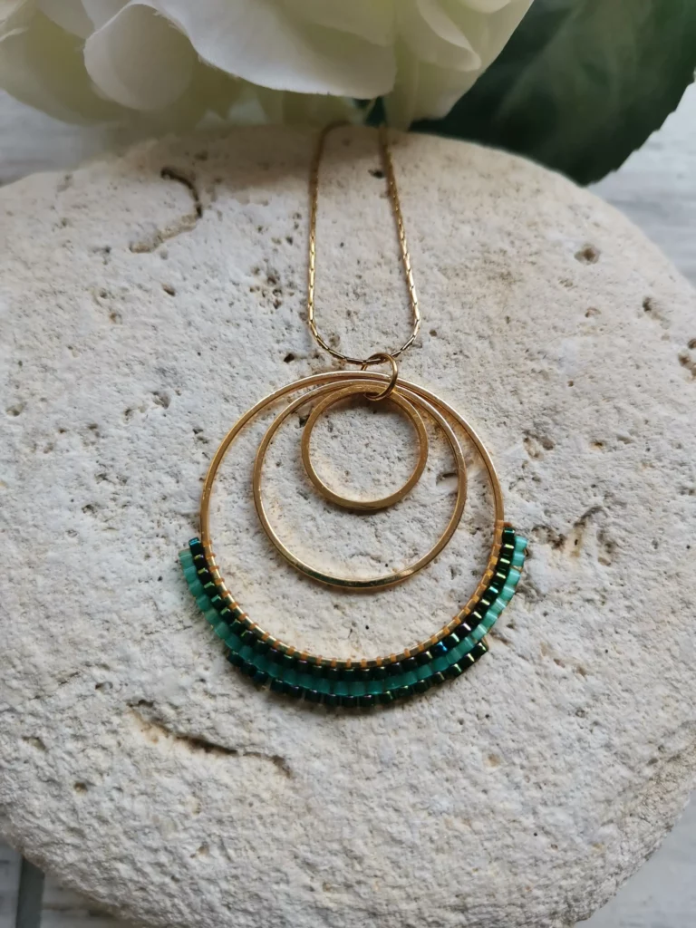 Collier avec 3 anneaux dorés de 3 tailles différentes, entrelacés. Le plus grand étant tissé en forme de 1/2 lune. La chaine est en acier inoxydable et le tout est posé sur un galet blanc.