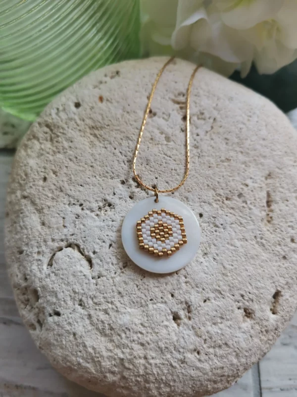 Collier tissage de perles Miyuki blanches et dorées, en forme de nid d'abeilles, collé sur palet de nacre rond . Monté sur chaine en acier inoxydable, il est posé sur une pierre blanche.