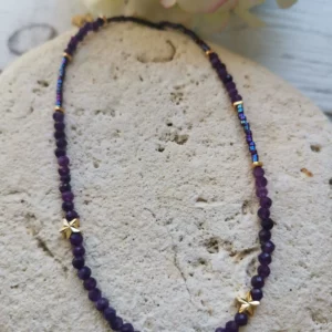 Collier tour de cou en perles d'améthyste de 3mm associées à des Miyuki violettes. 2 petites étoiles plaquées or sur le devant du collier. Il est posé sur une pierre blanche.