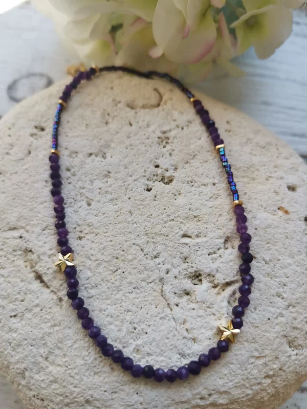 Collier tour de cou en perles d'améthyste de 3mm associées à des Miyuki violettes. 2 petites étoiles plaquées or sur le devant du collier. Il est posé sur une pierre blanche.